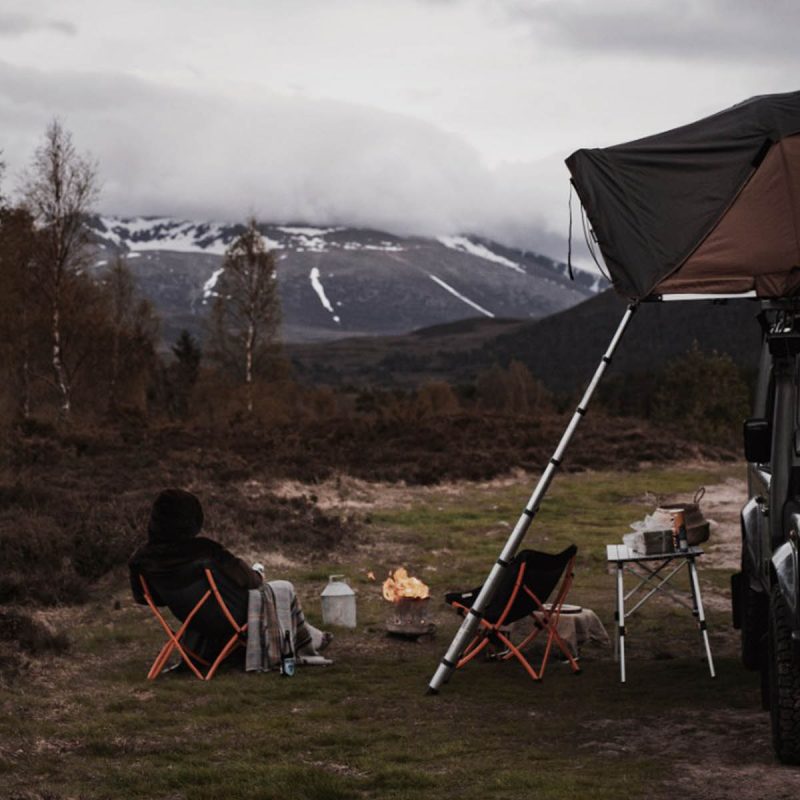 A person camping with their Land Rover with a roof tent and sitting by a camp fire.