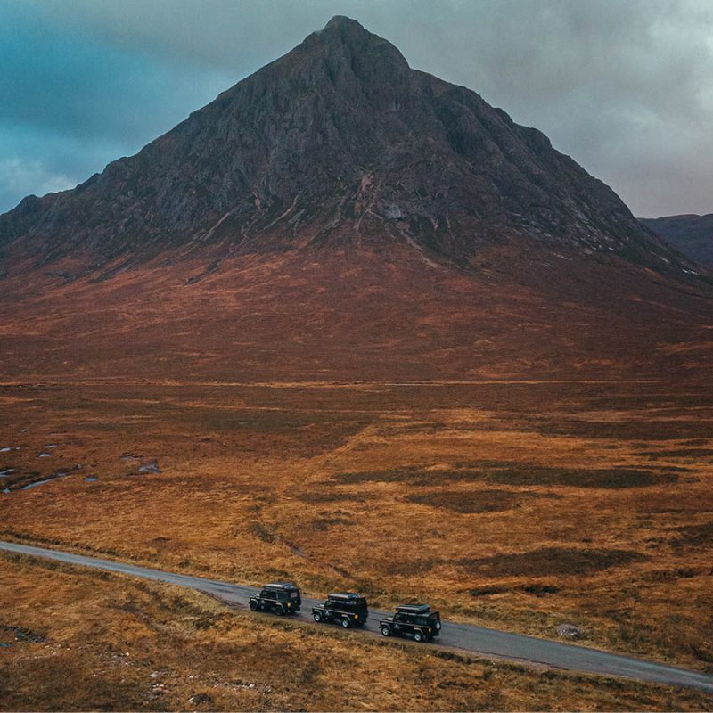 Three Nomadic Scotland Land Rover's driving across a rugged Scottish landscape with a peaked mountain in the background in Autumn.