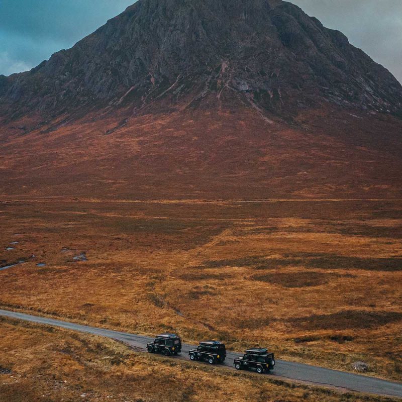 Three Nomadic Scotland Land Rover's driving across a rugged Scottish landscape with a peaked mountain in the background in Autumn.