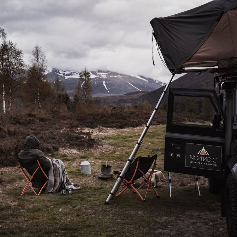 A person camping with their Land Rover with a roof tent and sitting by a camp fire.